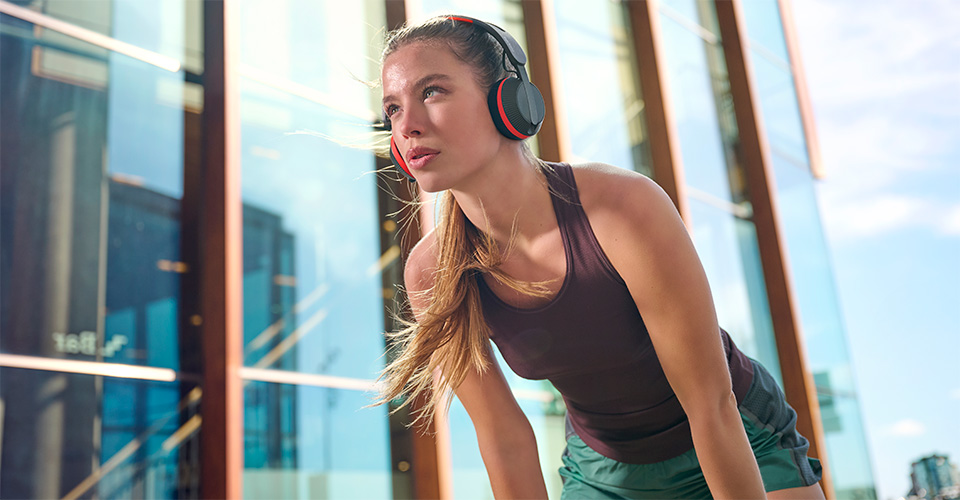 A&ccedil;ık havada egzersiz yaparken mola veren Philips A6219 kulaklık takan kadın, modern bir cam binanın &ouml;n&uuml;nde duruyor