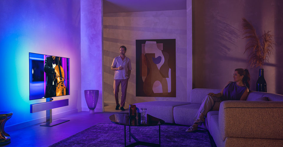 Şık bir oturma odasında Philips Ambilight TV'de &ccedil;ello performansı izleyen erkek ve kadın