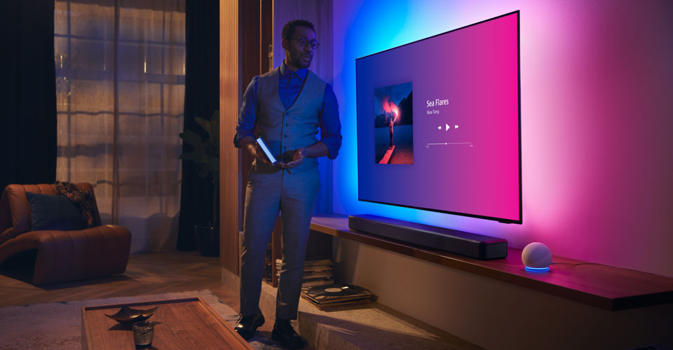 M&uuml;zik aray&uuml;z&uuml; ve Fidelio ses &ccedil;ubuğu bulunan Philips Ambilight TV'nin yakınındaki modern bir oturma odasındaki kişi