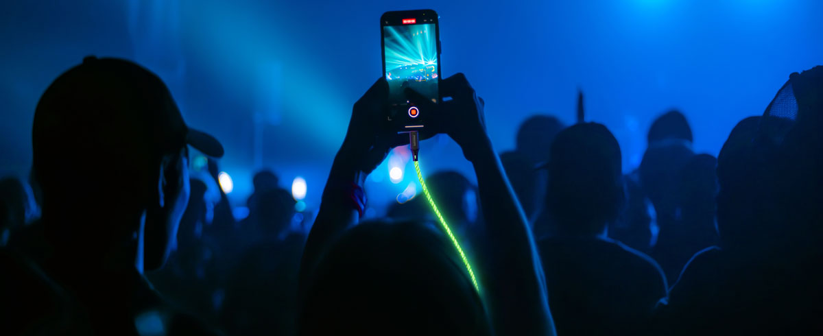 B&uuml;y&uuml;k bir kalabalığın &ouml;n&uuml;nde parlayan bir kabloya bağlı cep telefonunu yukarı kaldırıp etkinlikte anı yakalayan kişi